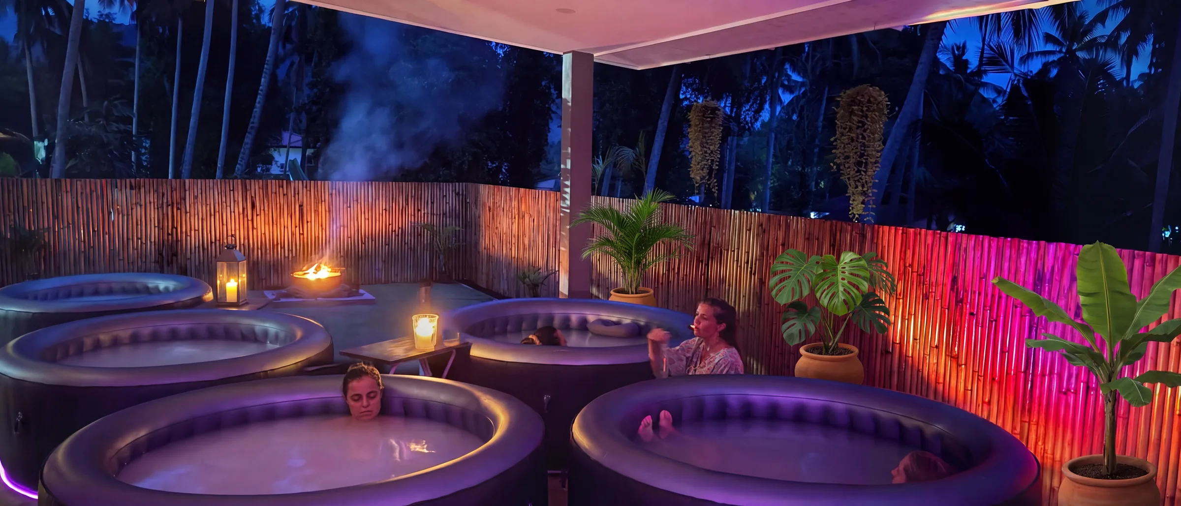 Sechs Pools unter dem Palmblattdach des Badehauses Bathtime am Abend, drei Gäste im Wasser, Feuerschale und Laterne — Foto, KI-erweitert