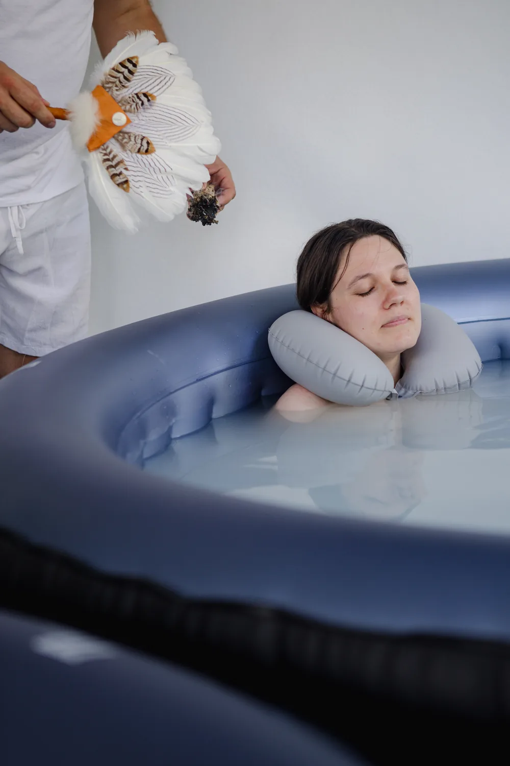 Ein Gast ruht im milchig-weißen Wasser eines Pools am Badehaus, eine Begleiterin hält eine Feder